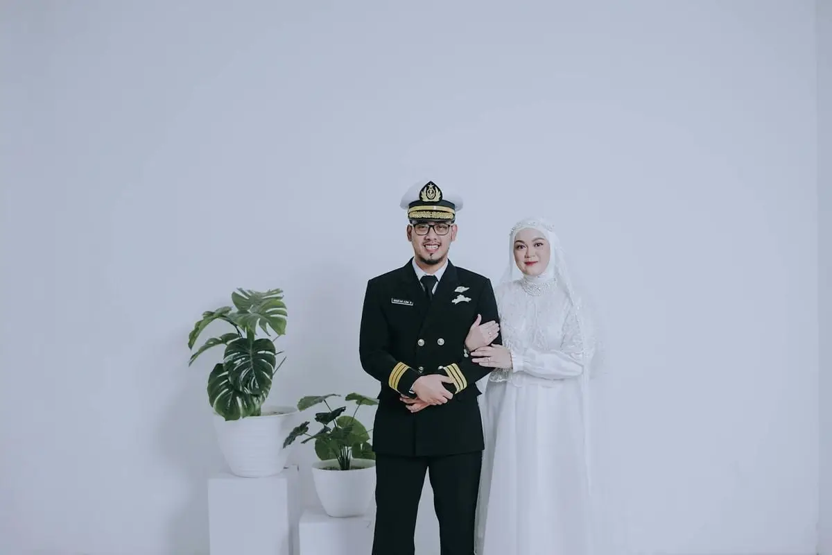 Bride and groom in military uniform and white gown, posing with potted plants.