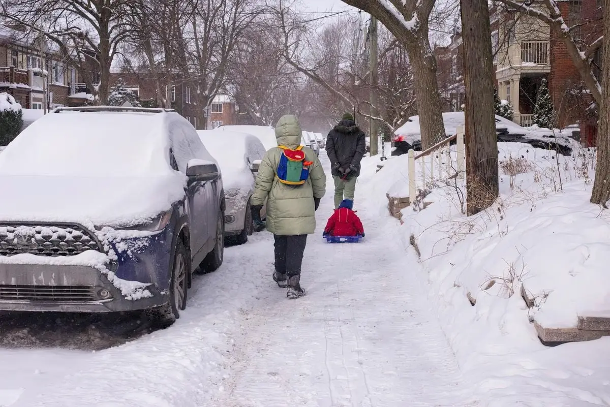Kids with backpacks navigate a snowy sidewalk, pulling a sled in a residential neighborhood.