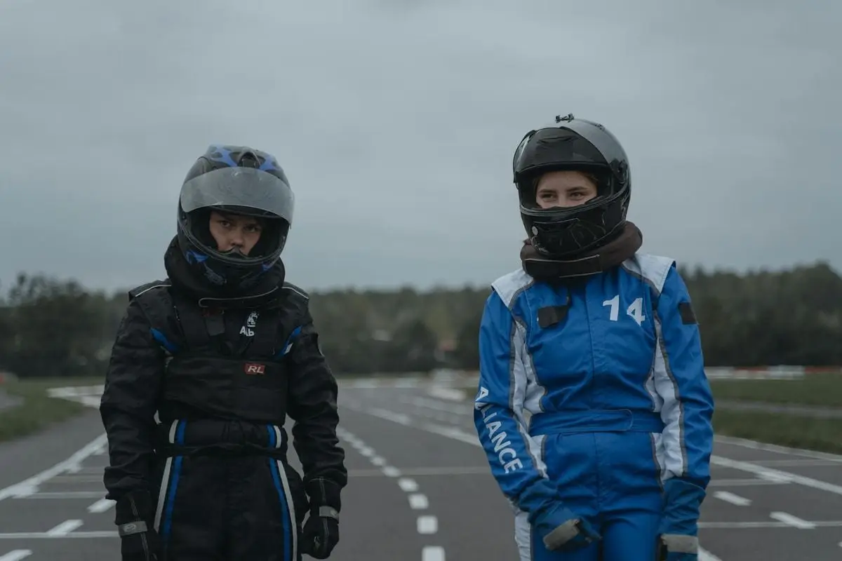 Two young drivers in racing suits and helmets standing on a go-kart track.