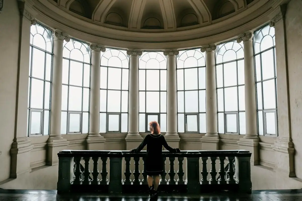 A woman gazes out from an elegant atrium featuring large arched windows and classic architecture.