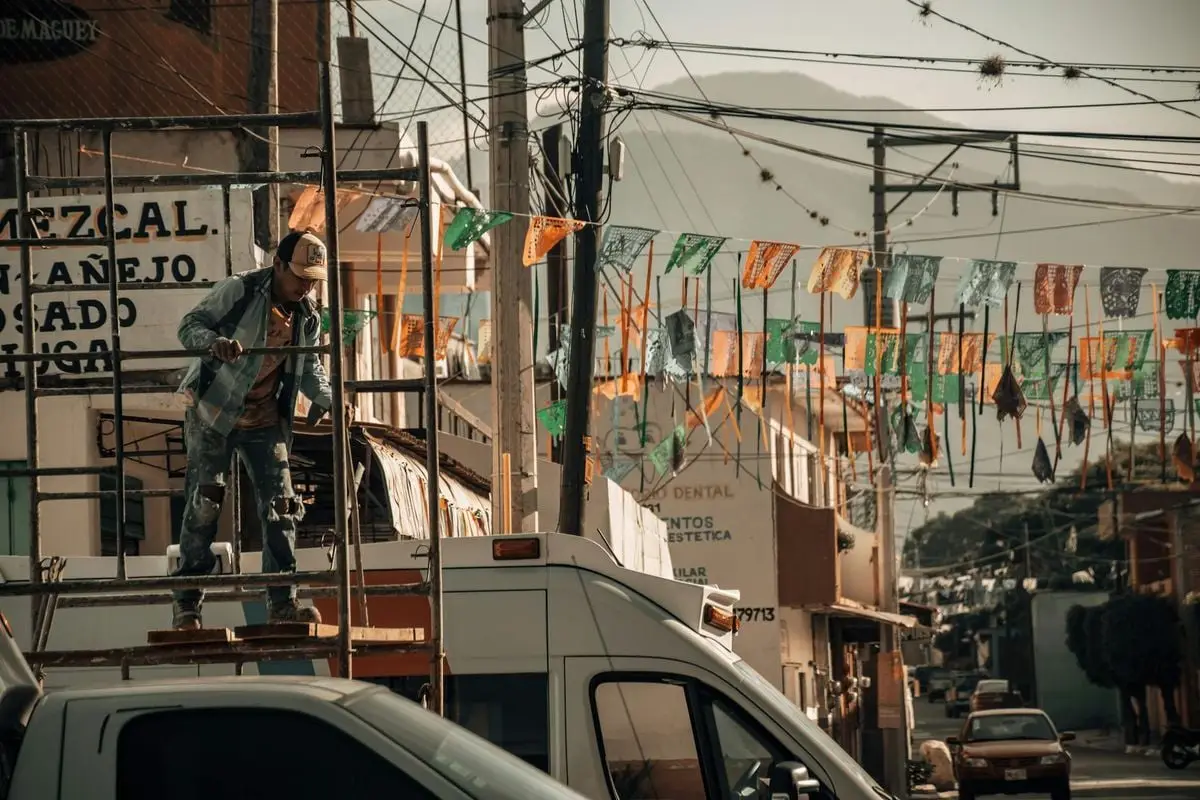 A construction worker stands on a scaffold in a vibrant urban area with colorful flags and cityscape.