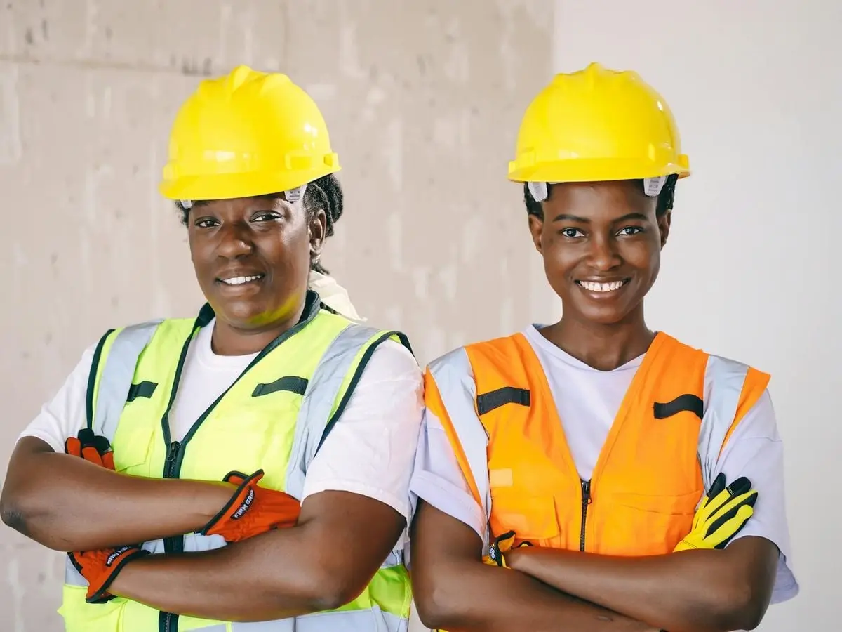 Two female engineers smiling confidently in hardhats and reflective vests, showcasing diversity.