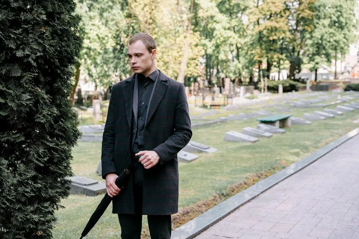 A man dressed in black holding an umbrella stands in a tranquil cemetery, evoking a sense of grief and remembrance.