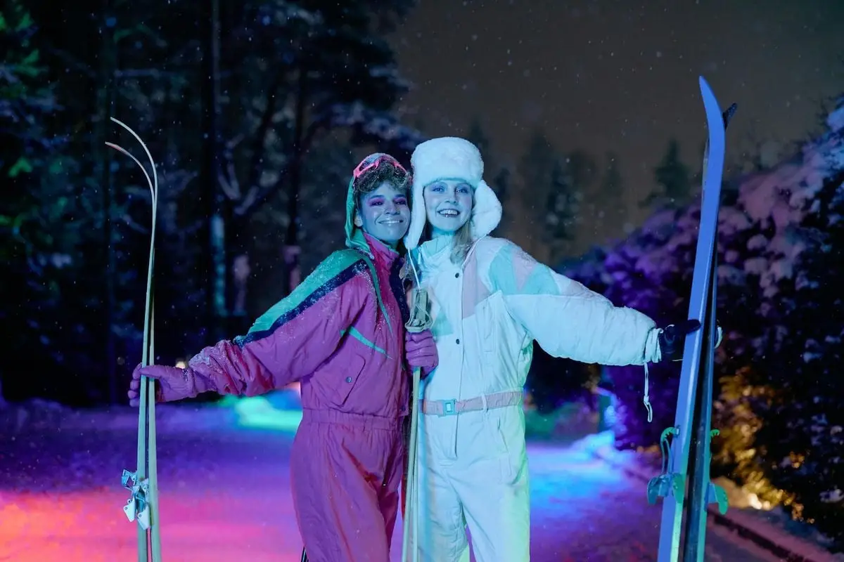 Two women enjoy night skiing under vibrant neon lights, showcasing stylish winter fashion.