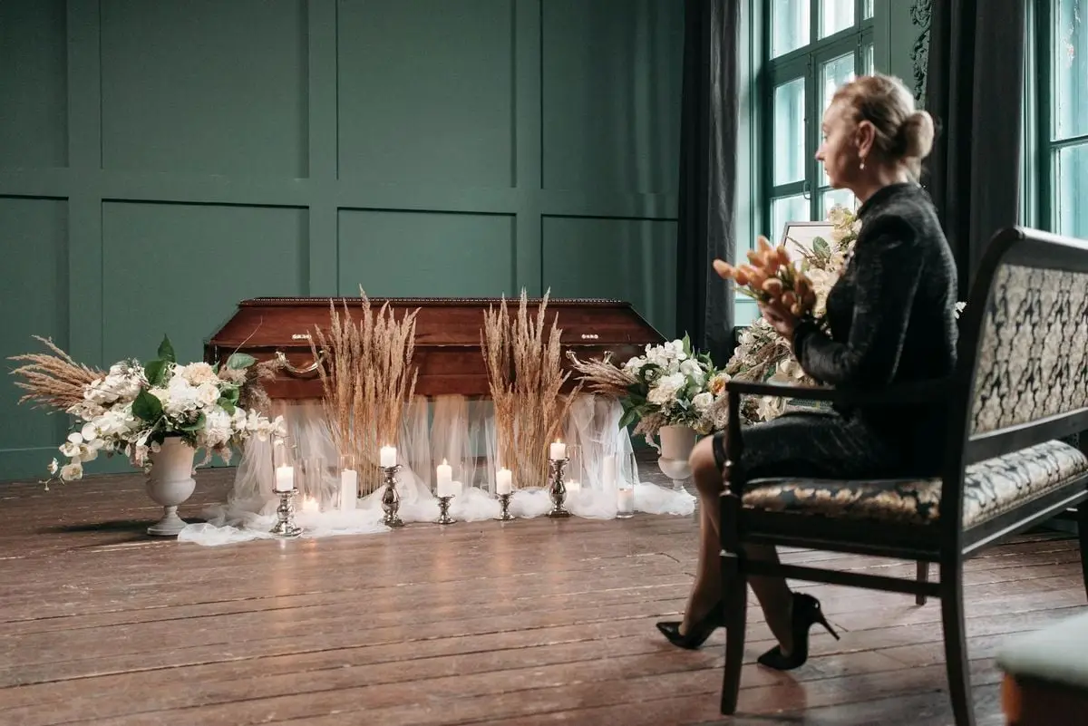 An indoor funeral scene featuring a coffin with floral arrangements and a mourning woman.
