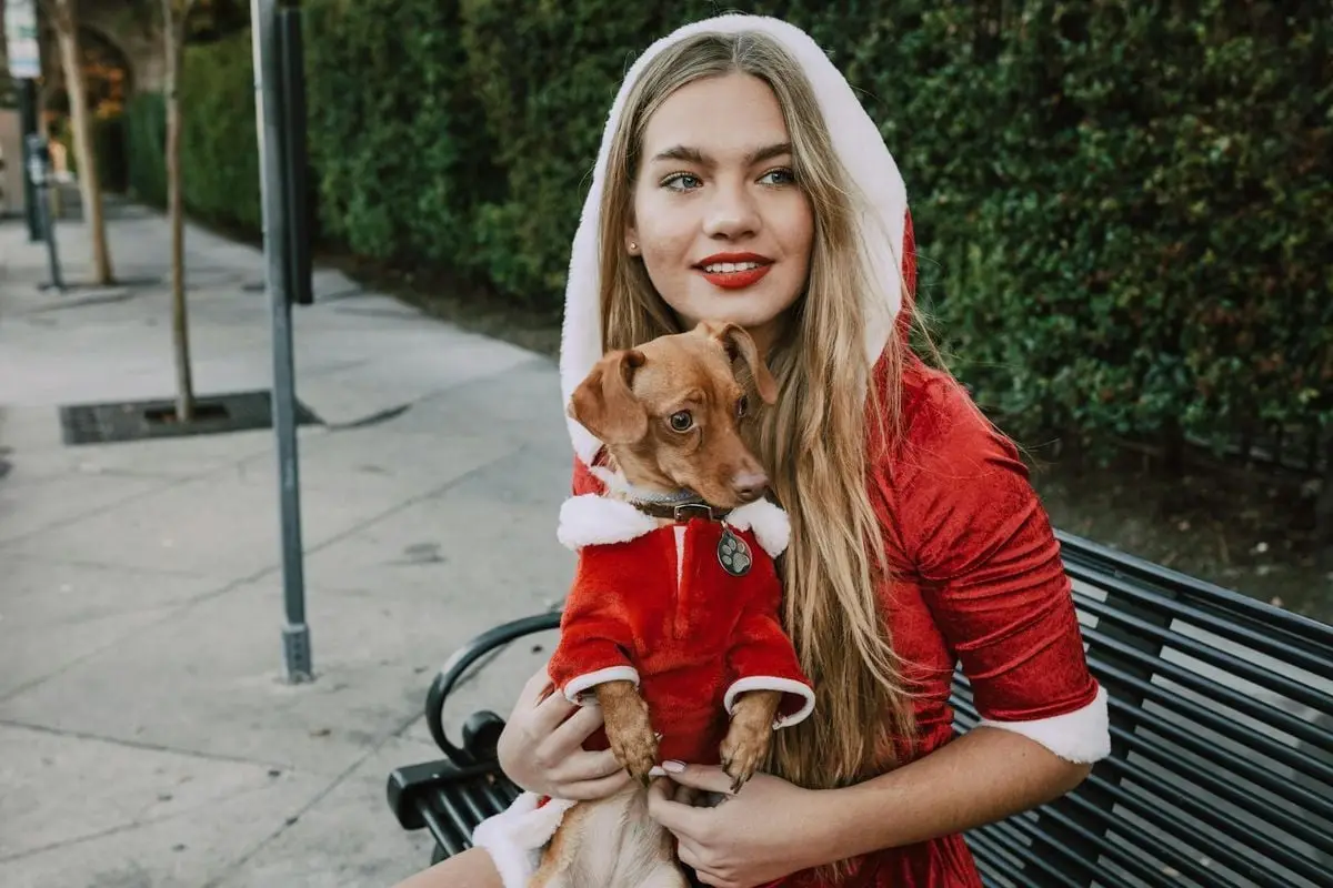 Young woman dressed as Santa holds a small dog in costume, seated on a bench outdoors.