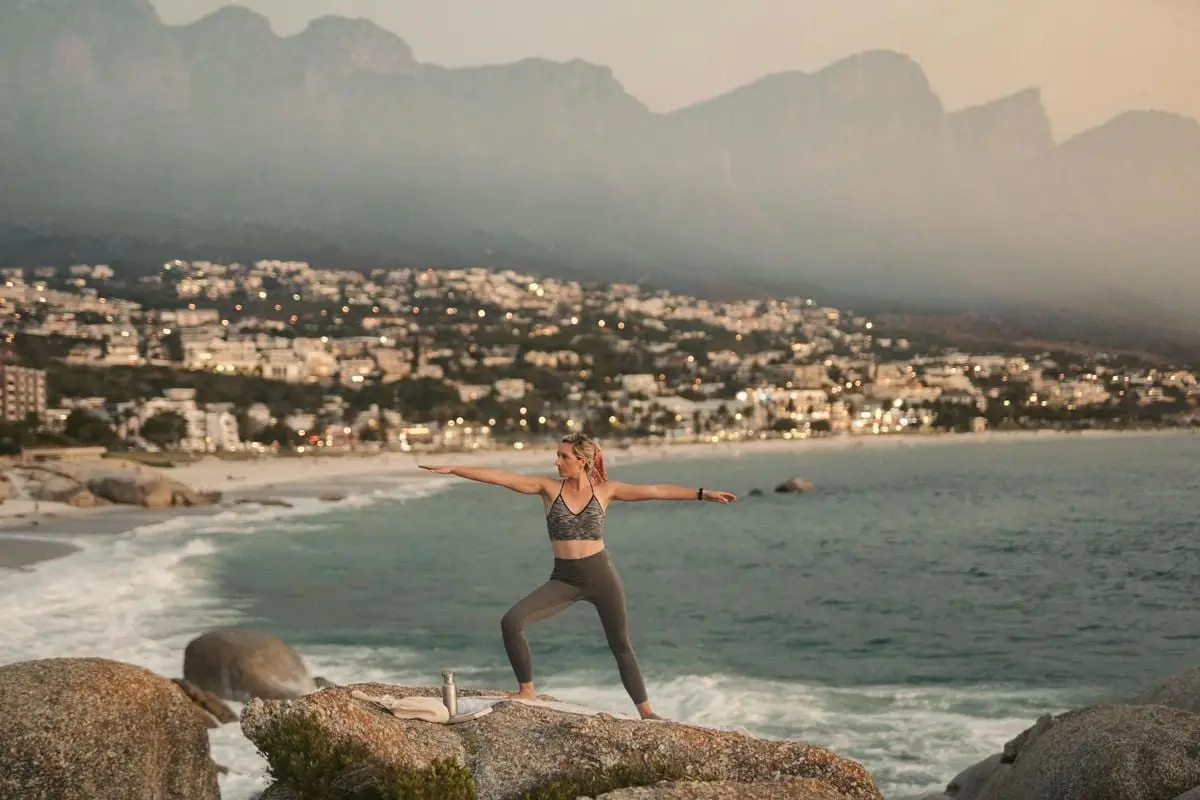 A woman performs the warrior pose on rocks overlooking the ocean at sunset in Cape Town, showcasing tranquility.