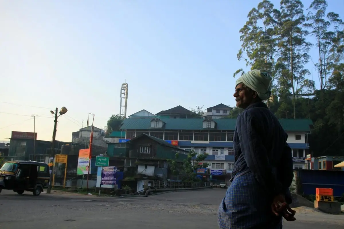 A South Asian man stands on a street in Munnar, India, with urban buildings and tall trees in the background.