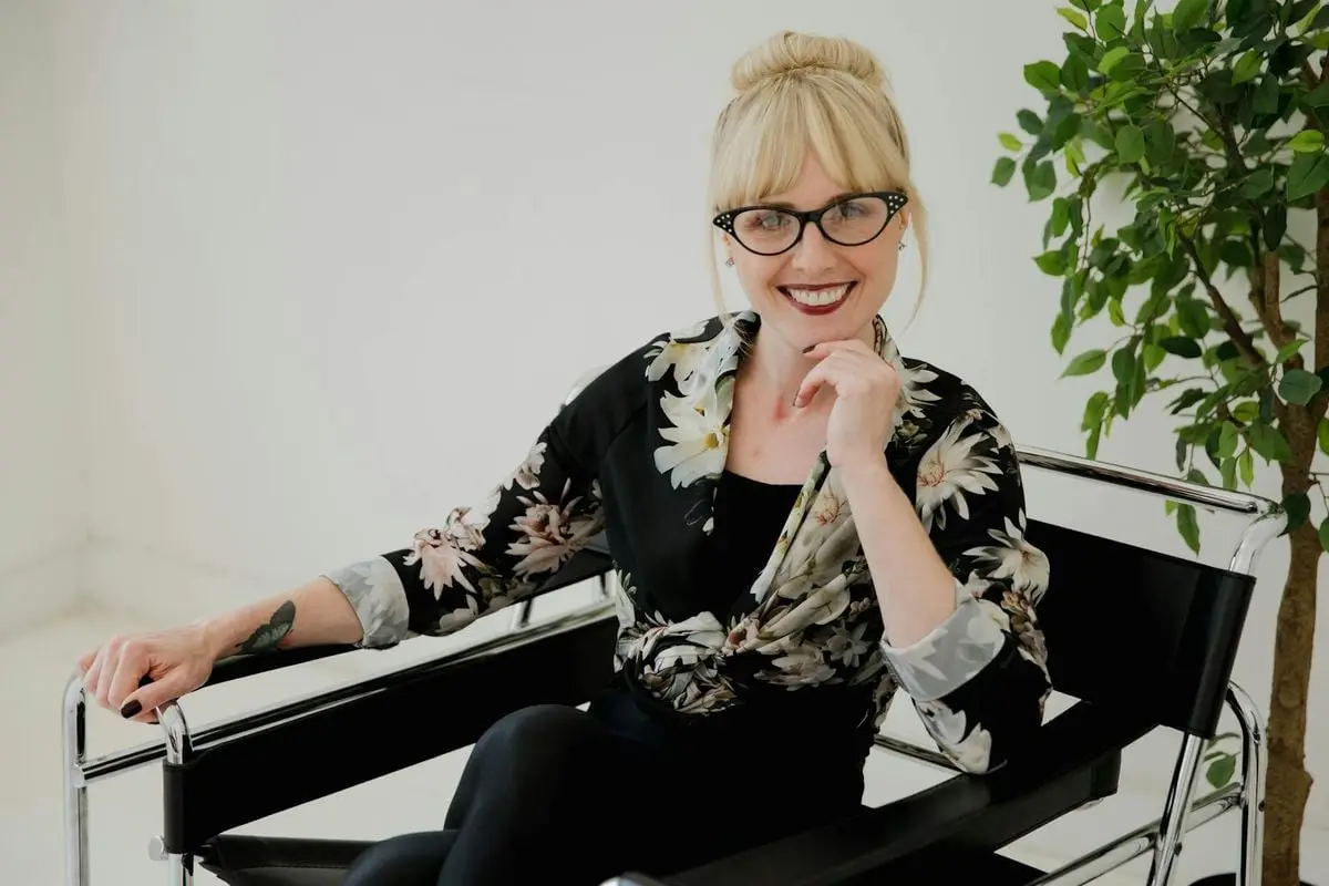 Blonde woman with eyewear smiling and sitting with hand on chin in a floral shirt, indoors.
