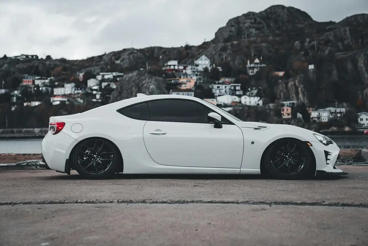 A white sports car parked by a lake with a scenic mountain town backdrop.