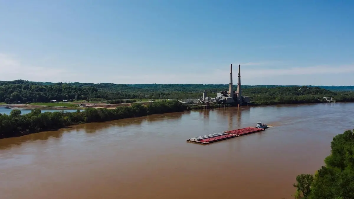 An aerial view showcases a barge on the Ohio River near Louisville with industrial buildings in the distance.