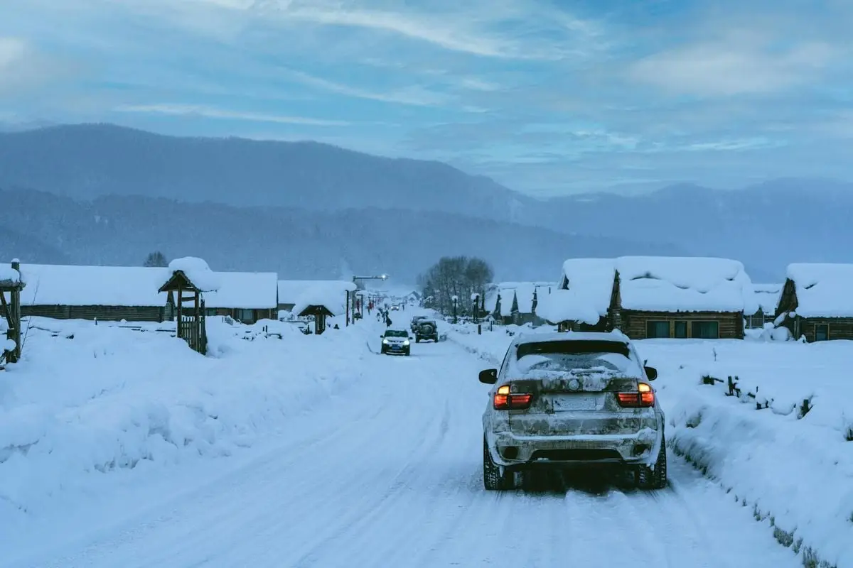 Snowy road lined with houses and moving cars against a mountainous backdrop on a winter day.