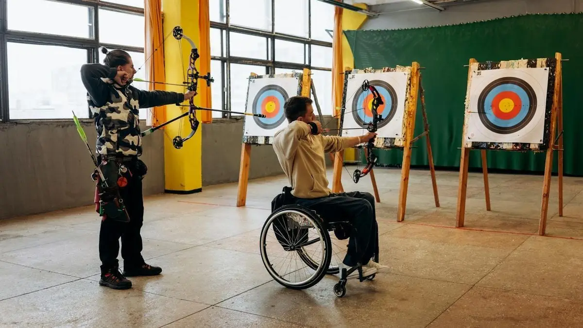 Two adults practicing archery indoors, one in a wheelchair, showcasing focus and determination.