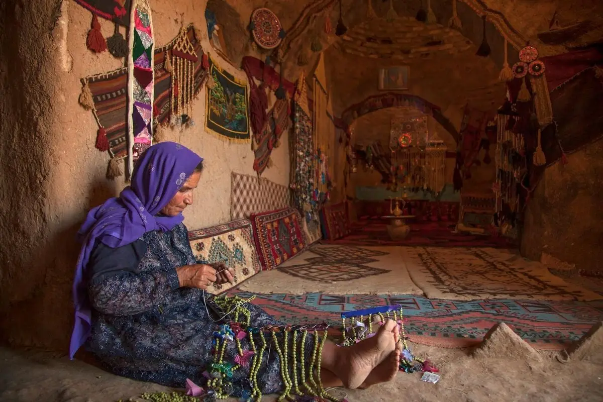 An elderly woman crafts colorful jewelry in a traditional Turkish interior, showcasing rich cultural decor.