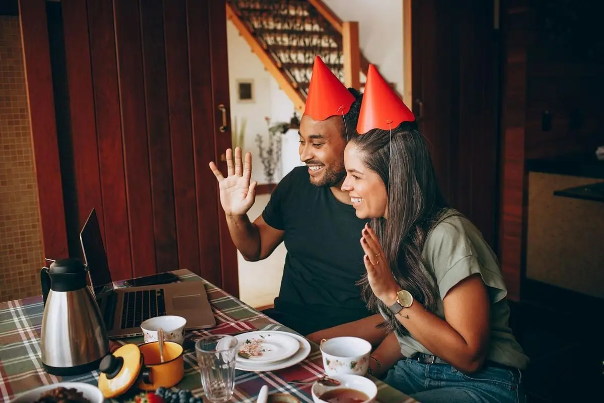Two adults celebrating a birthday via video call, wearing party hats and waving at a laptop indoors.