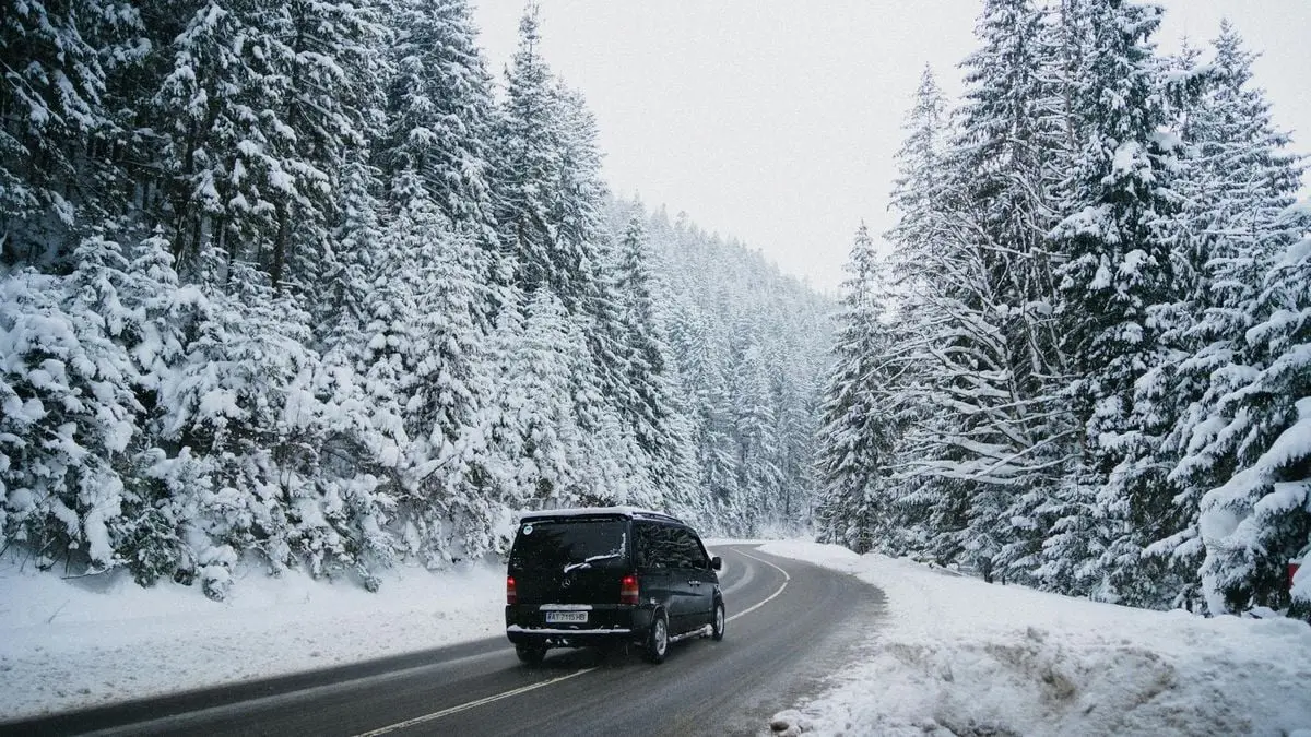 A black van driving on a snow-covered road through a winter forest with evergreen trees.