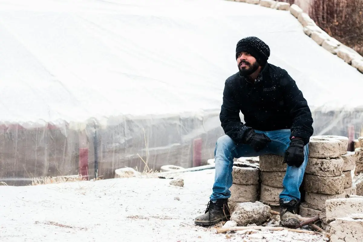 Man sitting on blocks in snowy outdoor setting, wearing winter clothing.