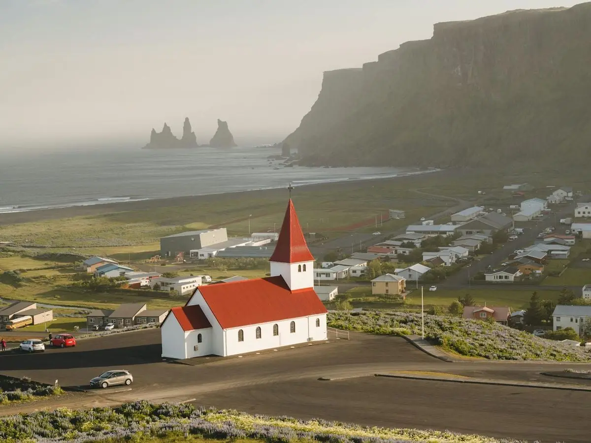 Aerial view of the iconic red-roofed Vik Church with Reynisdrangar sea stacks along the stunning Icelandic coastline.