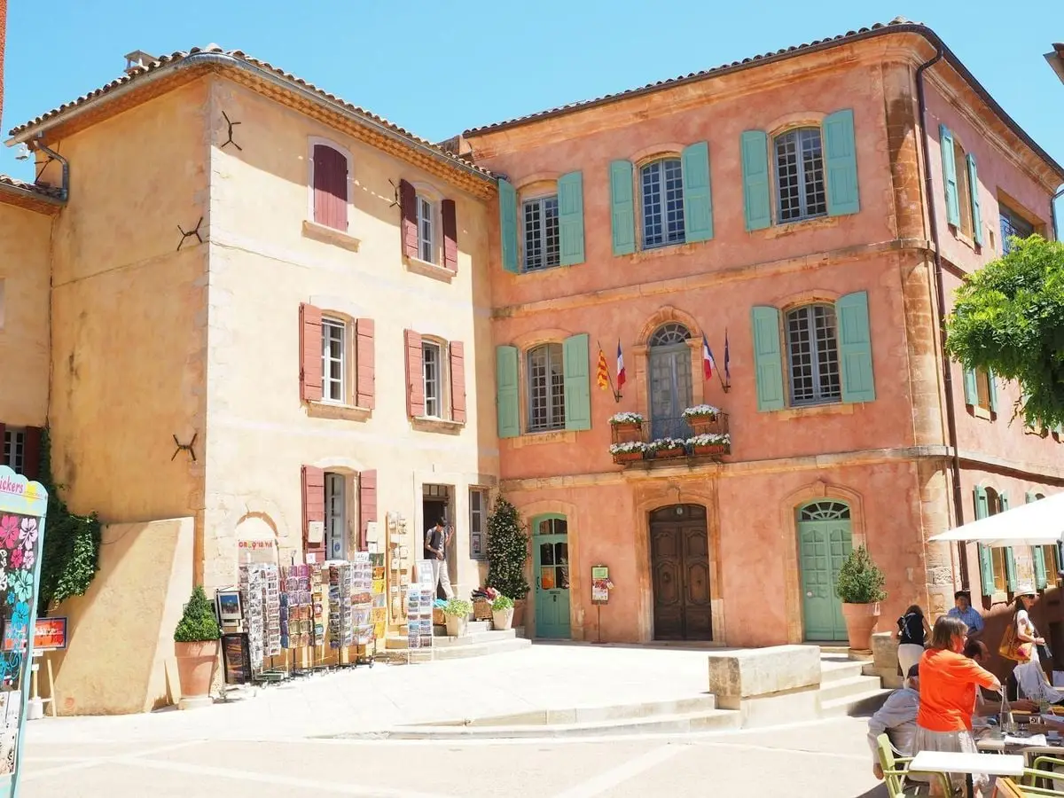 A sunny day in a picturesque French village square with colorful buildings and lively atmosphere.