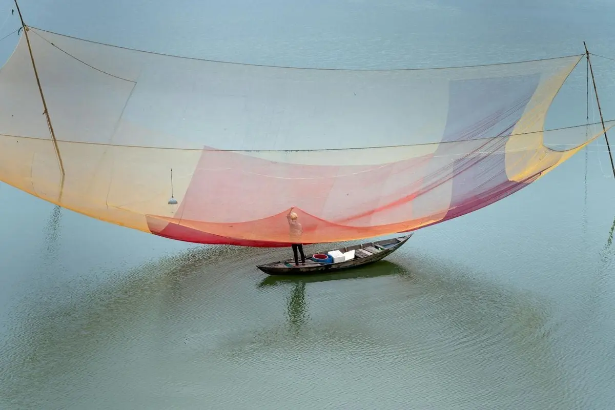 Aerial view of a fisherman on a boat with a large colorful fishing net cast on calm water.
