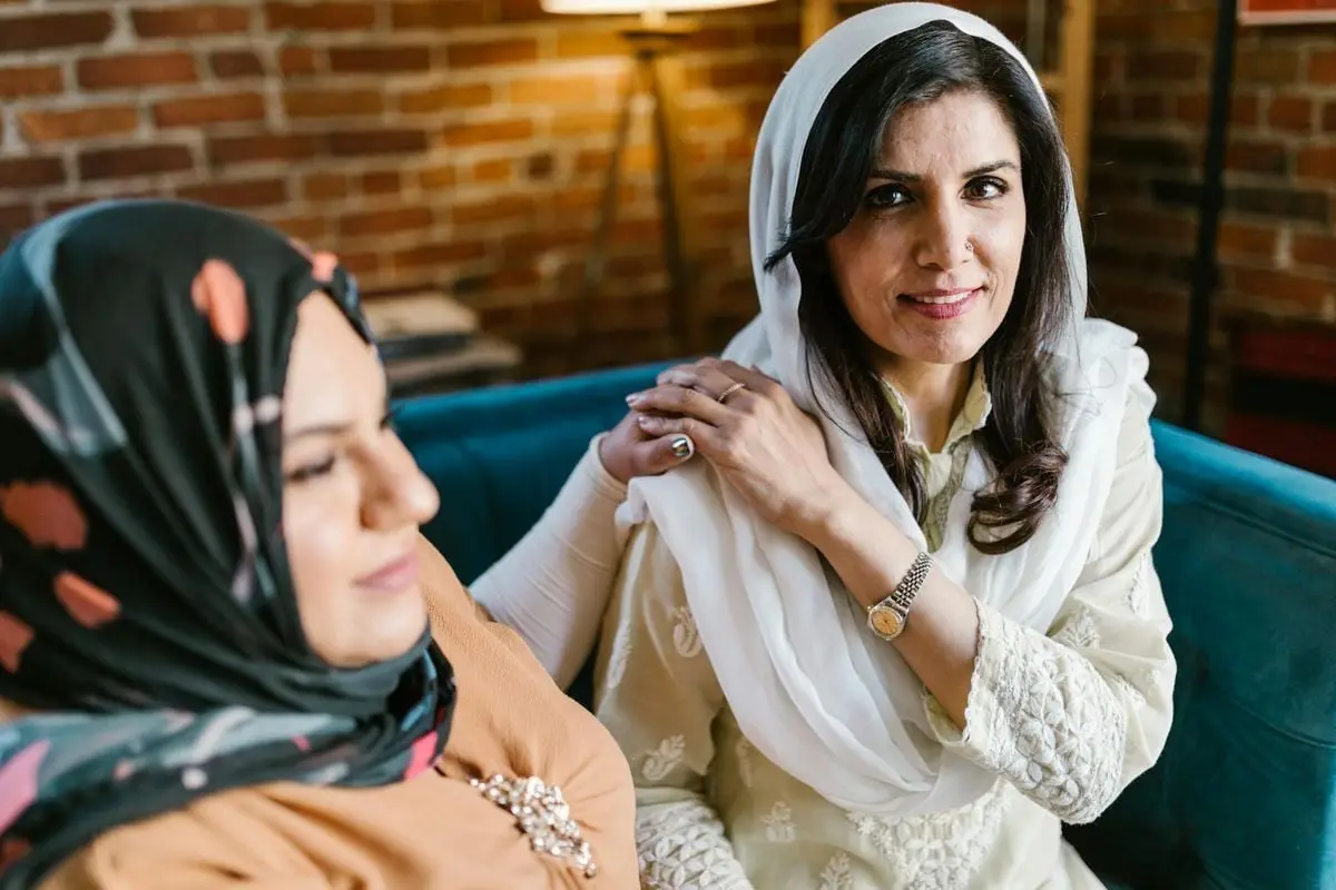 Two women in hijabs sit on a blue couch against a brick wall, sharing a moment of friendship.