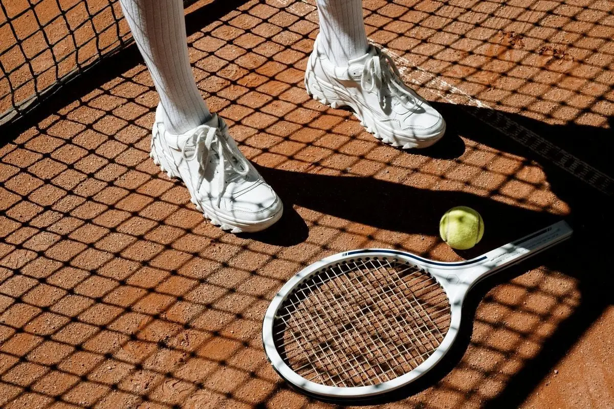 Tennis player's legs with racket and ball on clay court under bright sun and shadow patterns.