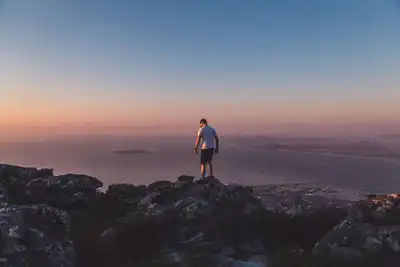 A lone hiker stands on a rocky mountain overlooking Cape Town at sunset.
