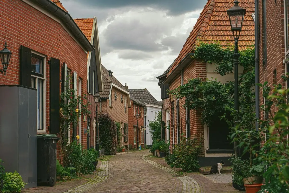 A picturesque European street with brick houses, cobblestone path, and lush greenery.