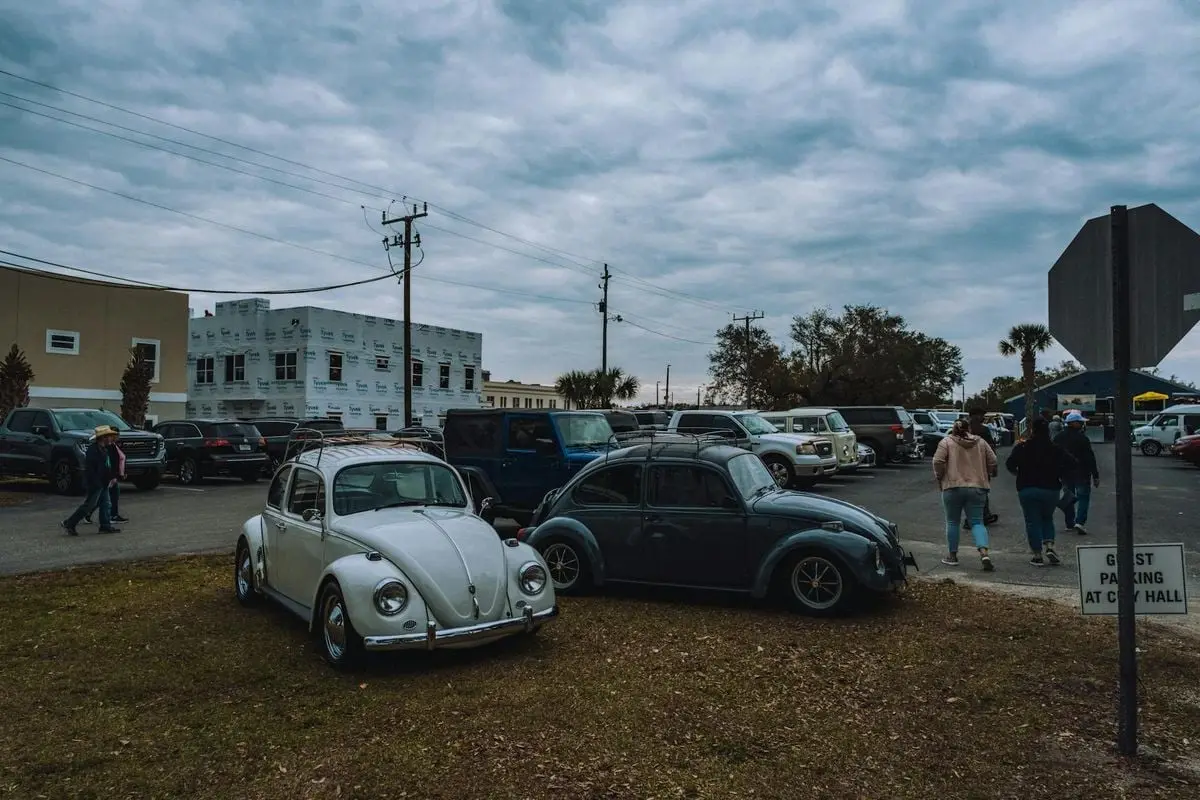 Two vintage Volkswagen Beetles parked in a town parking lot under cloudy skies with urban background.