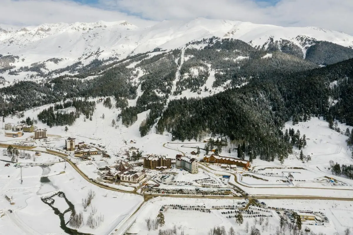 Drone shot of a snowy mountain ski resort with coniferous trees and snowy peaks.