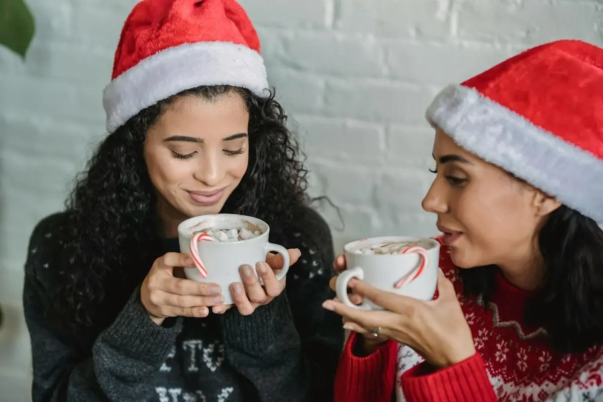 Two women in Santa hats enjoying hot chocolate with marshmallows indoors, evoking festive warmth.