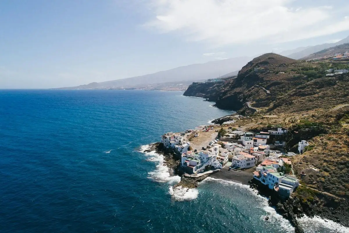 A picturesque aerial view of a coastal town nestled by a blue ocean and rugged cliffs under a bright sky.