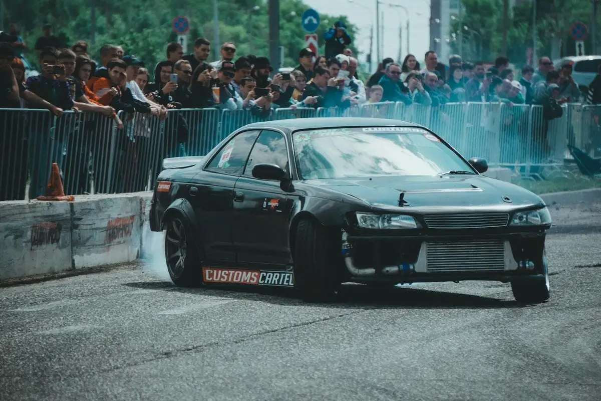 Black Toyota Mark II performs a drift maneuver at a crowded urban race event.