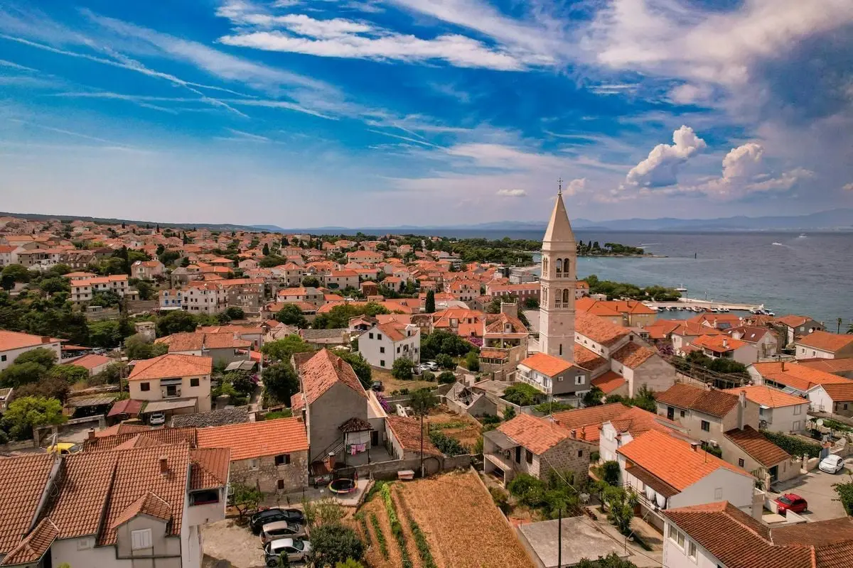 A picturesque aerial view of a coastal town with orange rooftops and church tower under a vibrant sky.