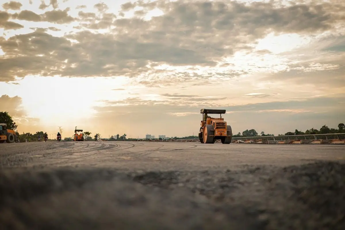 Steamroller on a road under construction with a scenic sunset backdrop and cloudy sky.
