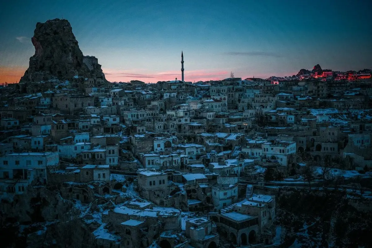 Aerial view of Cappadocia's ancient town under snow with towering rock formation at twilight.