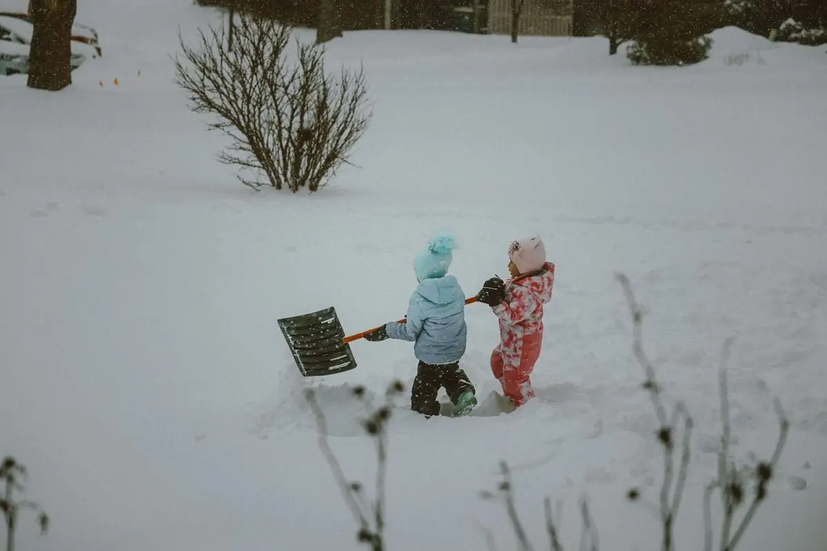 Two children in winter outfits play and shovel snow in a snowy landscape, enjoying winter fun.