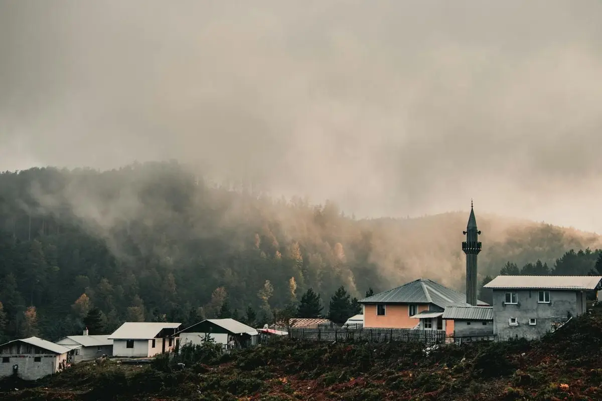 A serene mountain village shrouded in fog, featuring a mosque and forested landscape.