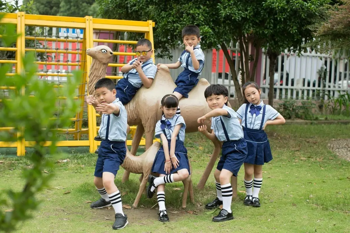 Children in uniforms pose on a camel statue on a school playground, showcasing playful school spirit.