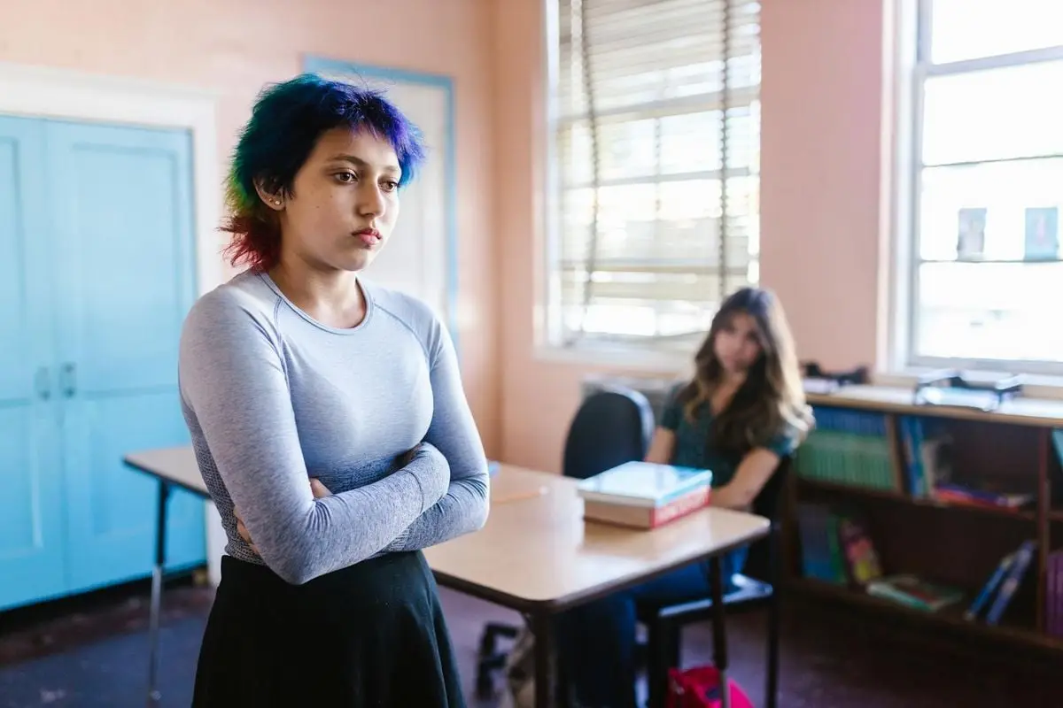 Student with vibrant hair crosses arms in school classroom, engaging and expressive.