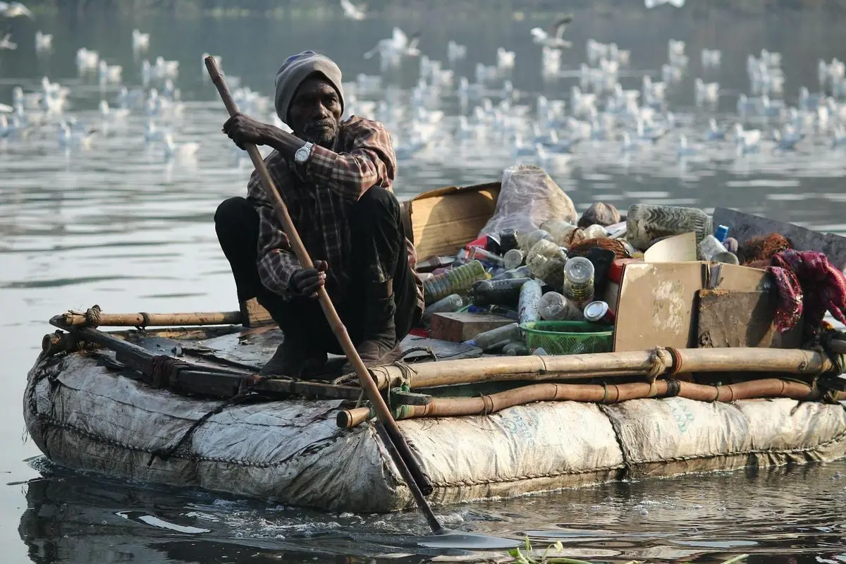 An adult man rows a makeshift raft filled with collected waste on a river, highlighting environmental cleanup.