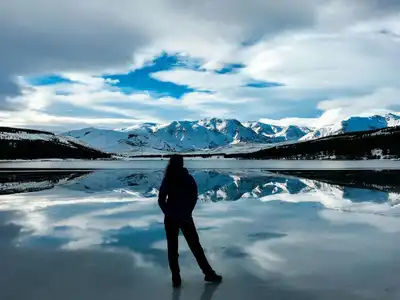 Back view of a woman standing on a frozen lake with reflections of snow-capped mountains in Patagonia, Argentina.