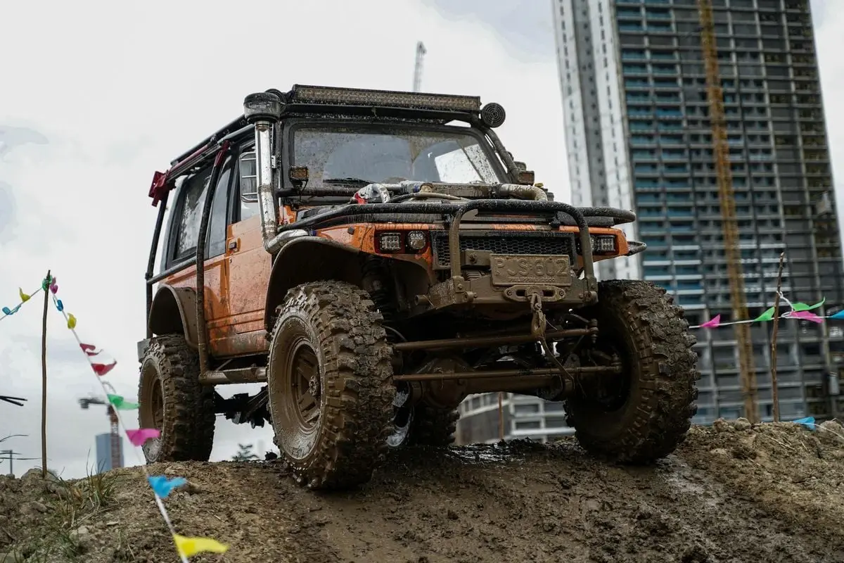 An off-road vehicle conquers a muddy hill with city buildings in the background.