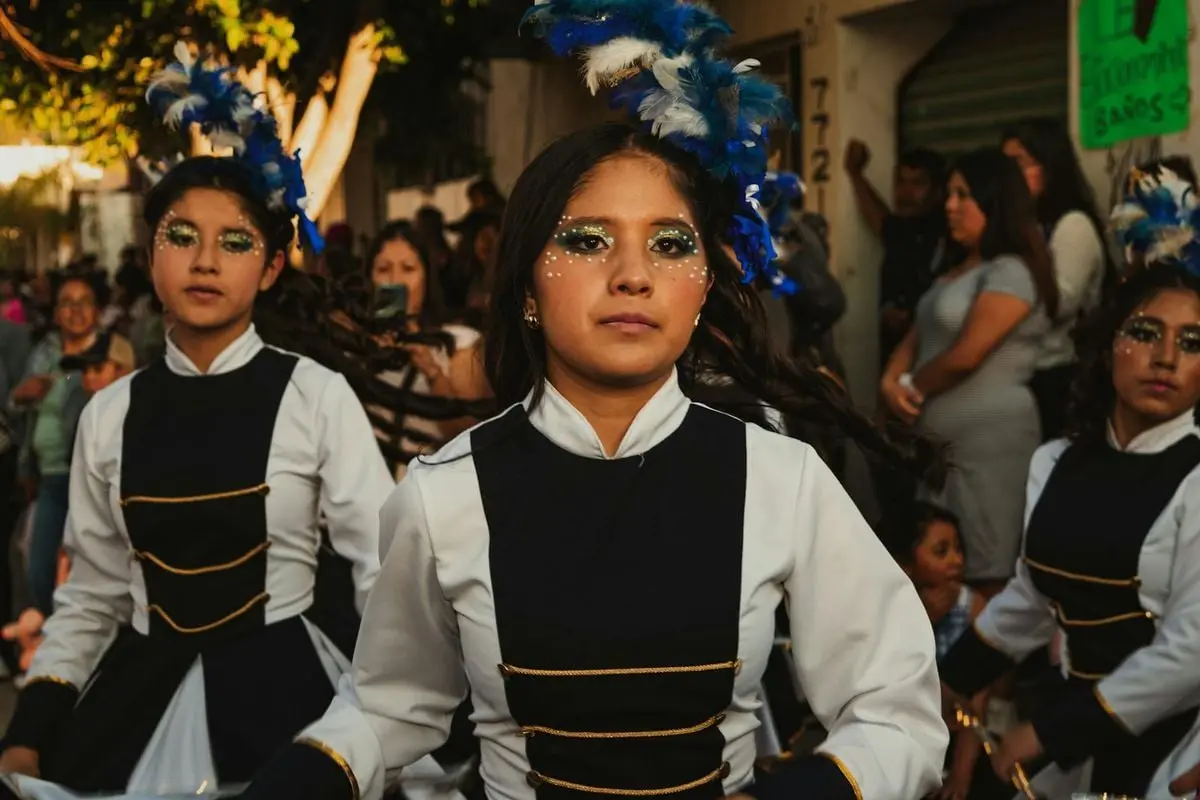 Three young women in traditional costumes participate in a lively street parade with colorful feathered headdresses.