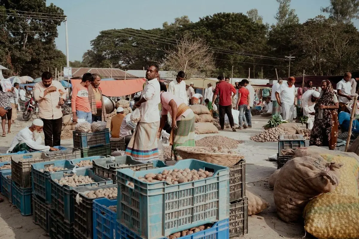 A vibrant outdoor market scene in Dhaka Division, Bangladesh, showing villagers trading potatoes and goods.