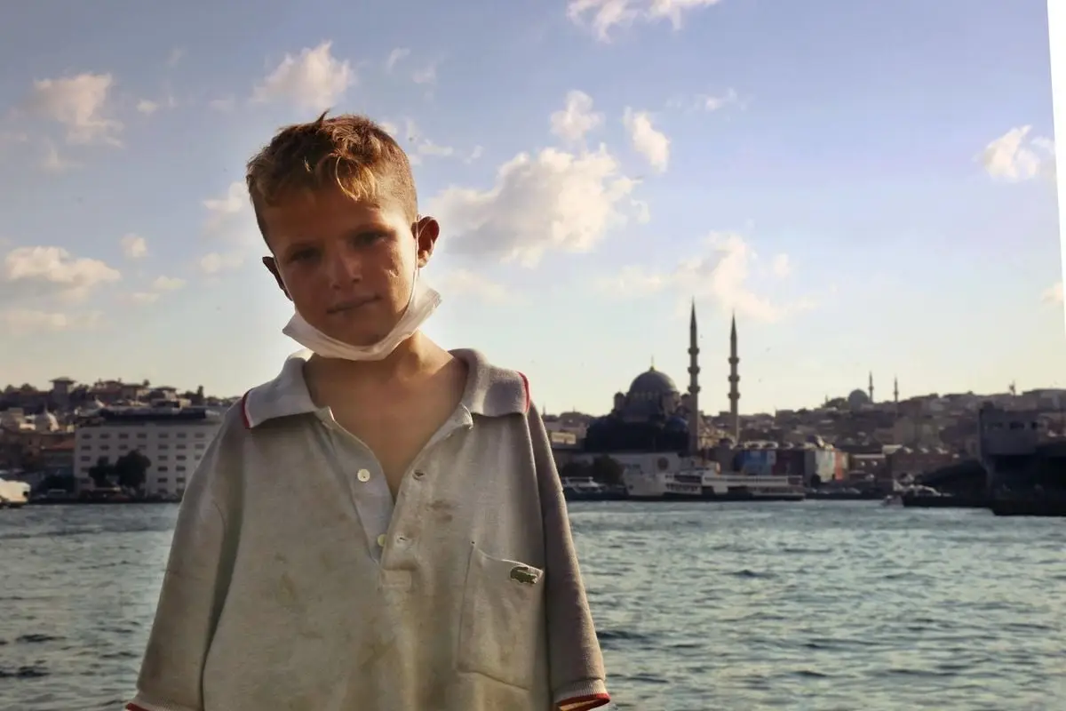 A young boy with a facemask poses by the Bosphorus Strait with Istanbul's skyline in the background.