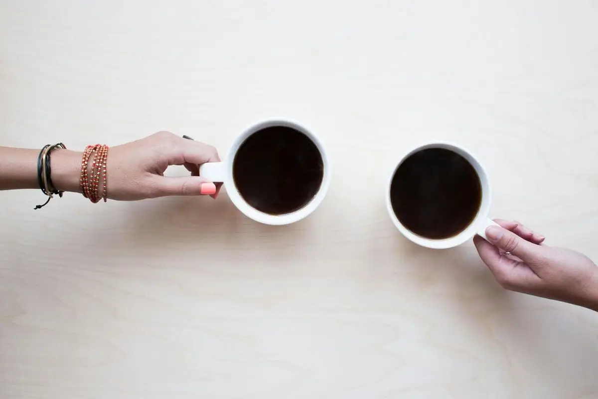 Two female hands holding coffee mugs on a light wooden table, symbolizing friendship and relaxation.
