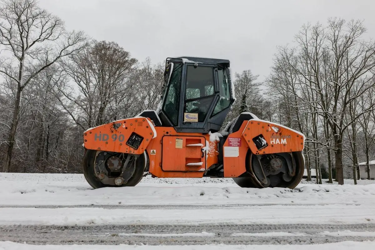 Bright orange tandem roller stands on snow-covered ground, surrounded by leafless trees in winter.