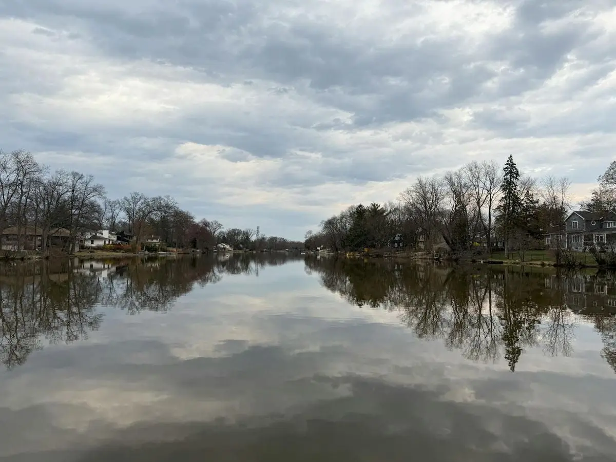 Calm lake surrounded by trees and houses reflecting a cloudy sky, capturing tranquil nature.