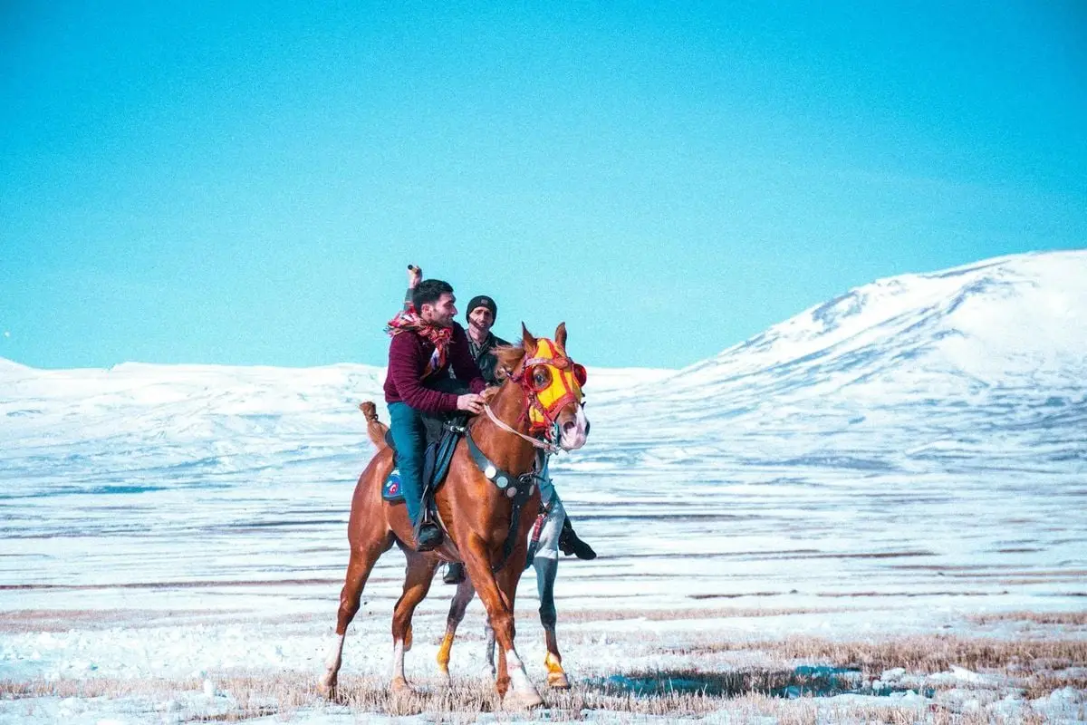 Two men horseback riding through the snowy plains of Kars, Türkiye in winter.